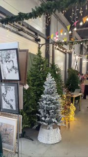 Вертикальный кадр с несколькими зелёными ёлками на выставочном стенде: гирлянды, картины и оформление торговой зоны на фоне.