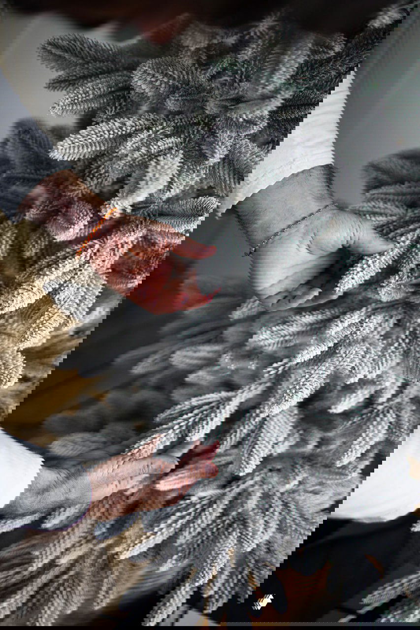 Крупный план: руки человека аккуратно расправляют заснеженные ветви искусственной ёлки «Донская заснеженная», детализация веток и текстура