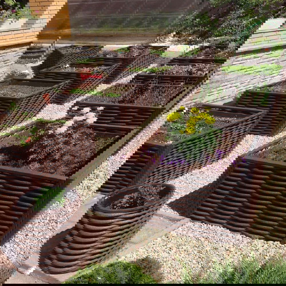Набор прямоугольных грядок и кашпо, обшитых полимерной лозой, на гравийной площадке у дома; демонстрация применения в огороде и цветоводстве.