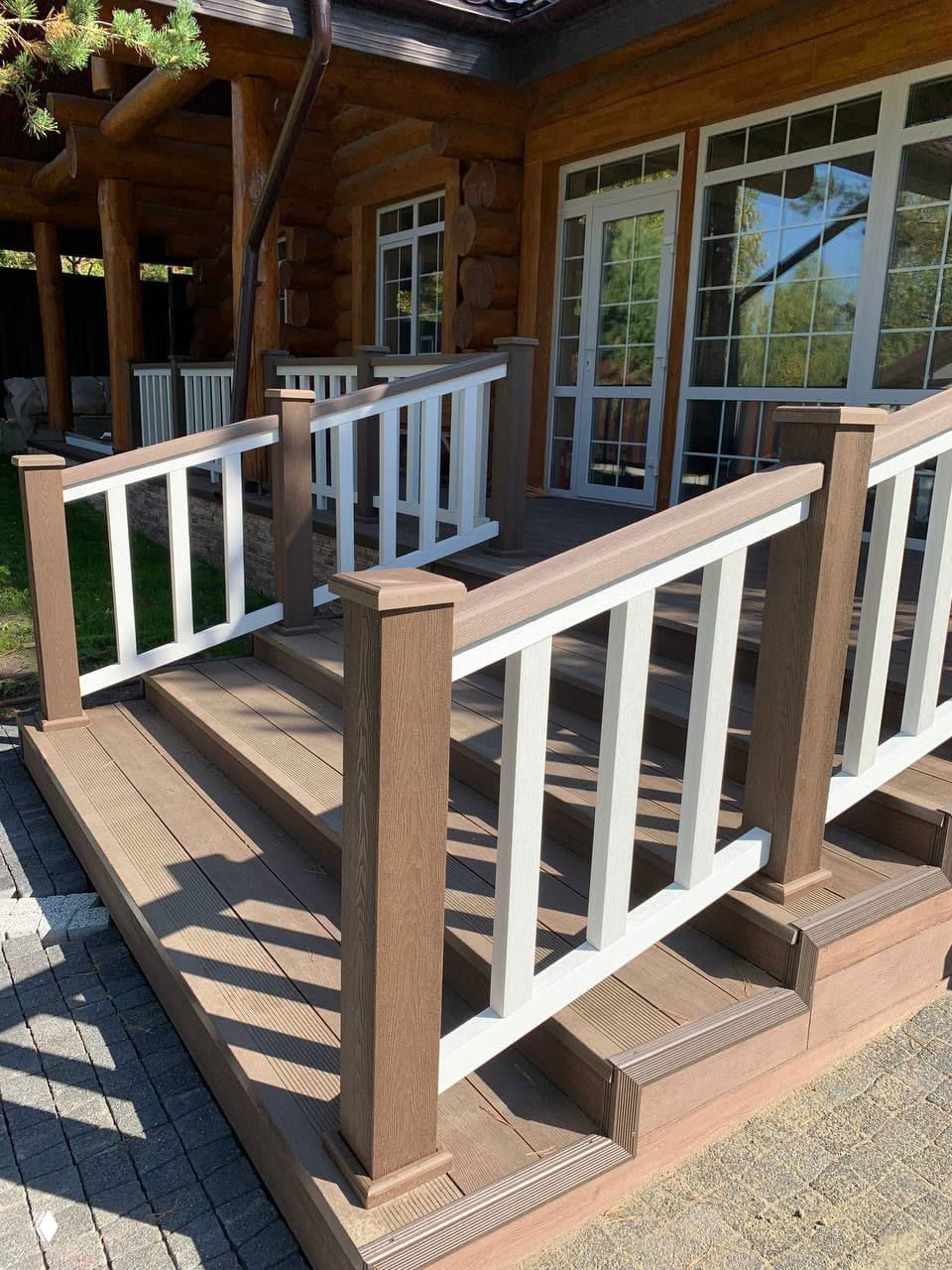 Уголовое фото композитной террасы: вид с лестницы, коричневые доски, белые вертикальные балясины и приглушённые тени от окружающих деревьев на настиле.