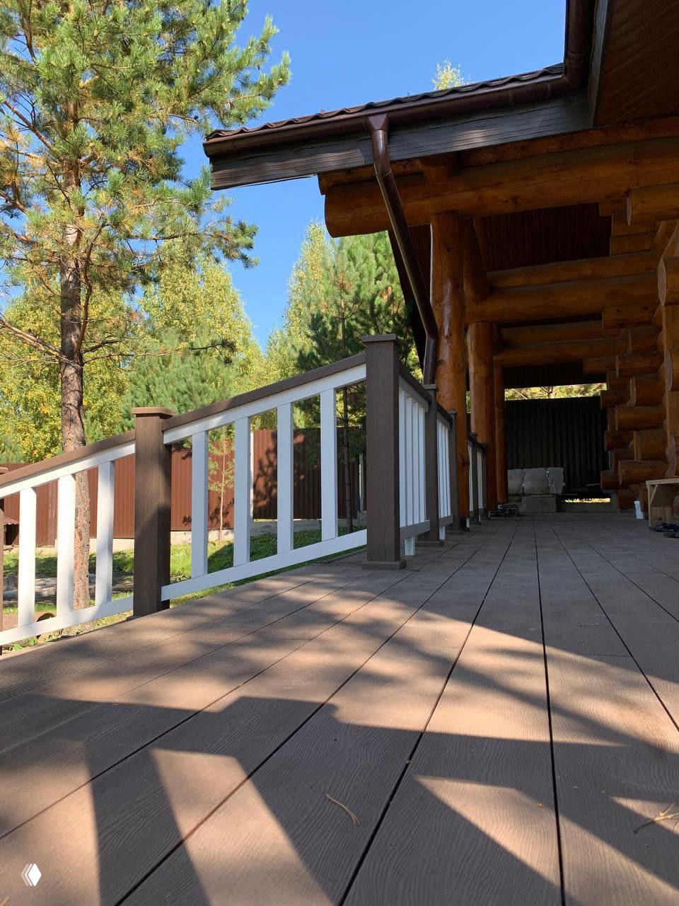 Перспектива настила и ограждения: ровная композитная доска, белые элементы перил, вдали хвойные деревья и чистое голубое небо над участком.