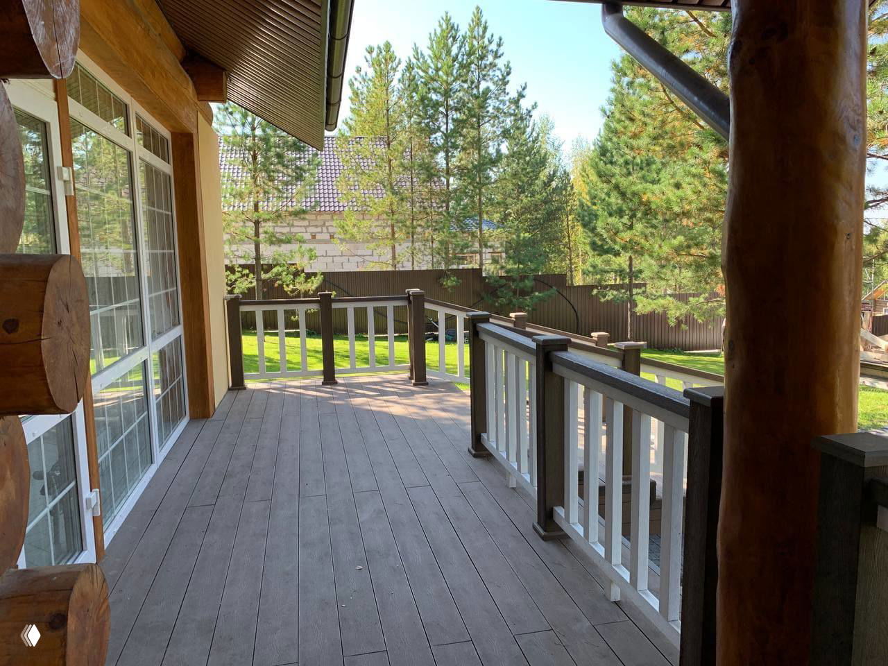 Вид на просторную веранду из ДПК у рубленого дома: ровный коричневый настил, ограждение с белыми стойками и зелёный сад на заднем плане в солнечный день.