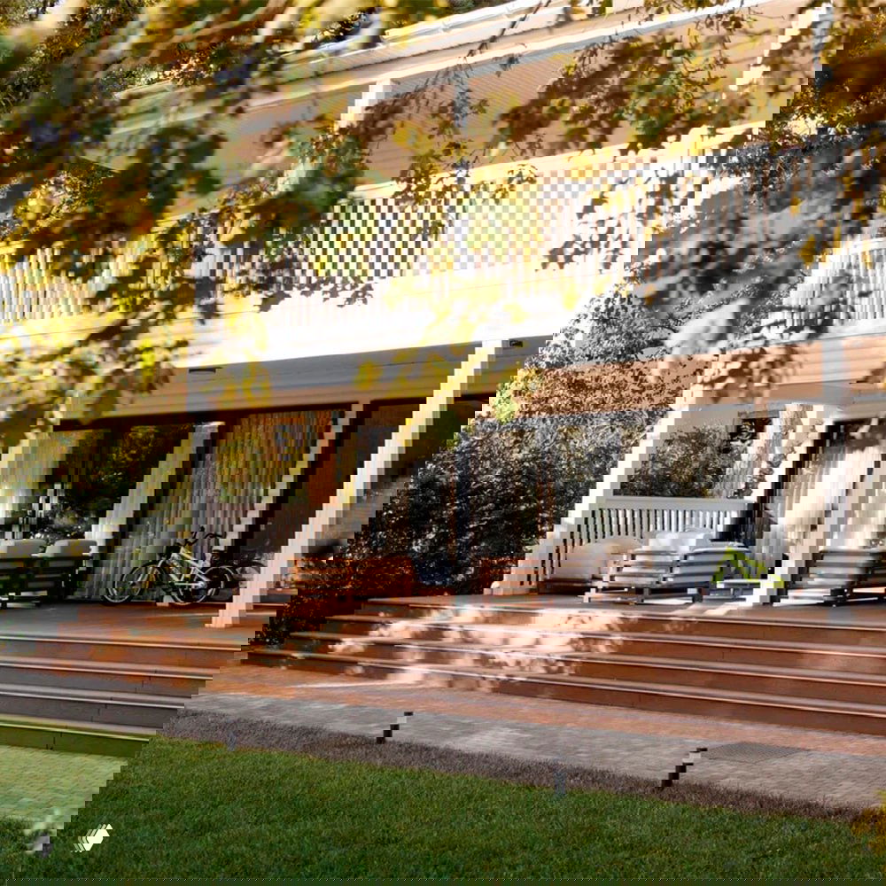 Веранда с настилом из ДПК: коричневый композитный настил у дома, листья деревьев на переднем плане, уютная внешняя зона отдыха.