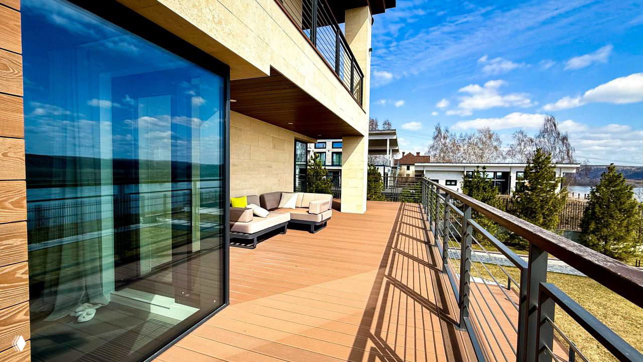 Угловая зона террасы из ДПК с диваном и стеклянной дверью: коричневые доски, ограждение и вид на озеленённый участок в солнечный день.