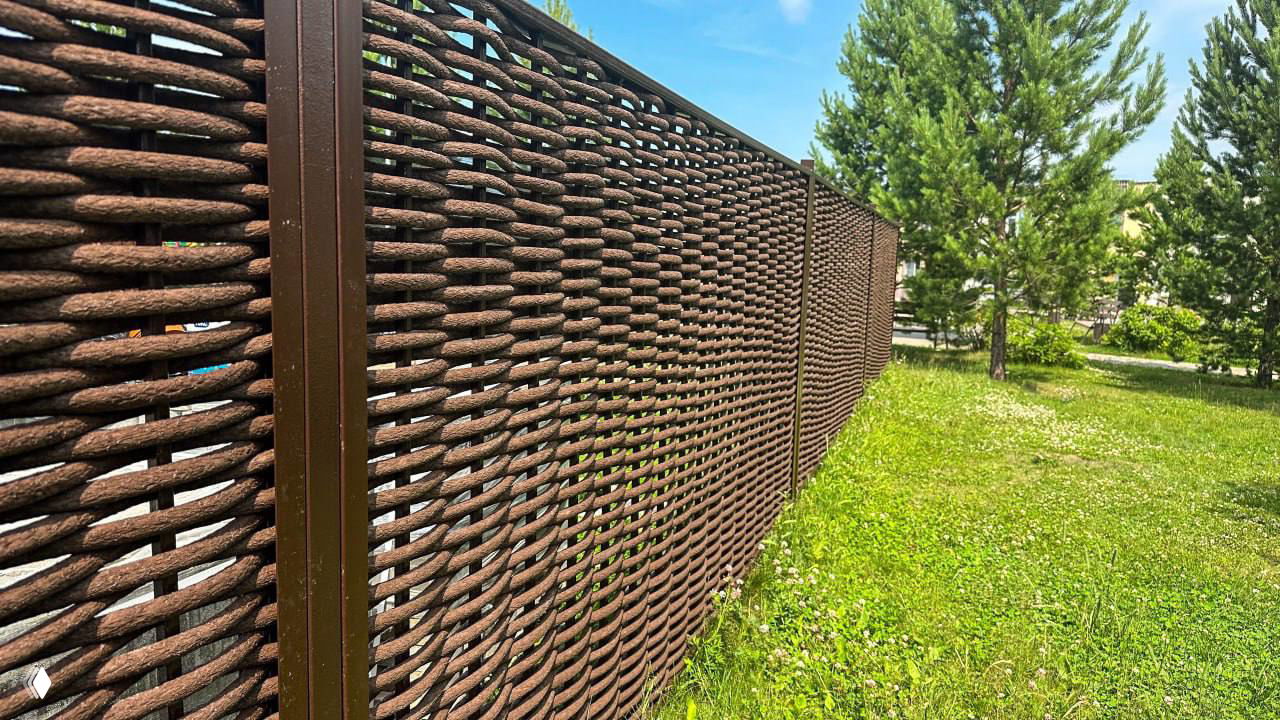 Крупный план полимерной лозы-забора: плетёная структура, коричневый цвет и металлические столбы на фоне газона.