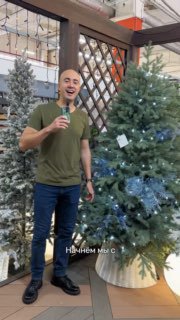 Мужчина улыбается и держит микрофон у украшенной искусственной ёлки в торговом помещении со стеллажами на заднем плане.