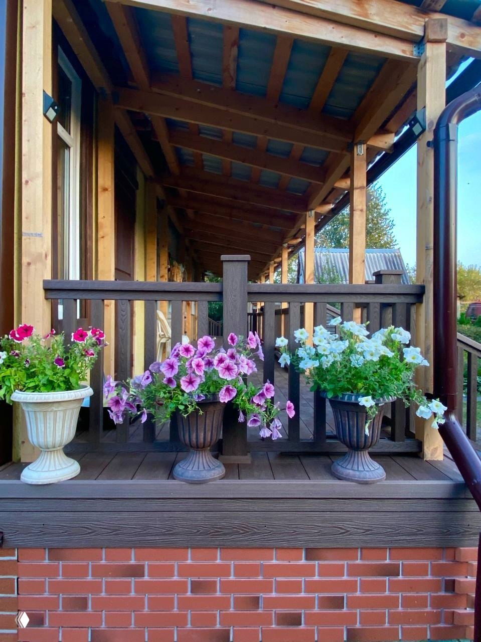 Терраса под навесом с перилами из ДПК, горшки с цветами на ограждении и деревянная конструкция в летней обстановке.