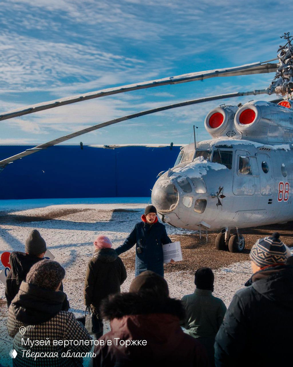 Экскурсовод ведёт группу туристов перед экспонатом — небольшим вертолётом в музее вертолётов в Торжке; снежная поверхность и голубое небо.
