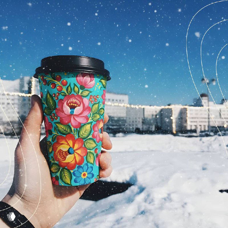 Рука в варежке держит расписную кружку с цветочным узором на фоне снежного поля и синего неба, вид крупным планом
