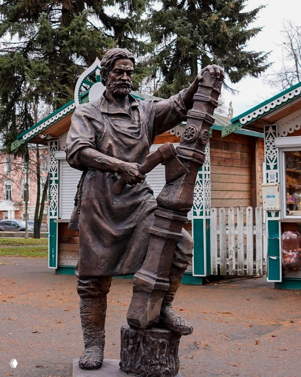 Бронзовая статуя ремесленника с большим инструментом — скульптура «Зодчий» на фоне вологодских деревянных киосков и елок