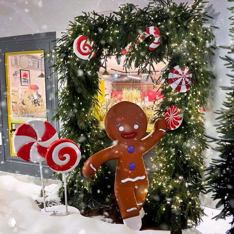 Праздничная уличная инсталляция с фигурой пряничного человечка, большими леденцами и гирляндой на фоне рождественского оформления городской площади или базара.