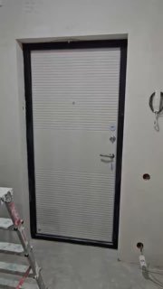 Входная металлическая дверь в черновом проёме: видна стена без отделки, стремянка, оголённые розетки и домофон — сцена замера.