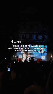 Сцена фестиваля вечером: артисты и ведущие на сцене перед публикой с телефонами, подсветка сцены и крупная проекция с текстом событий.