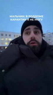 Кадр из вертикального видео: мужчина в тёплой шапке и куртке стоит на улице перед зданием, в кадре видна подпись о роддоме и карантине.