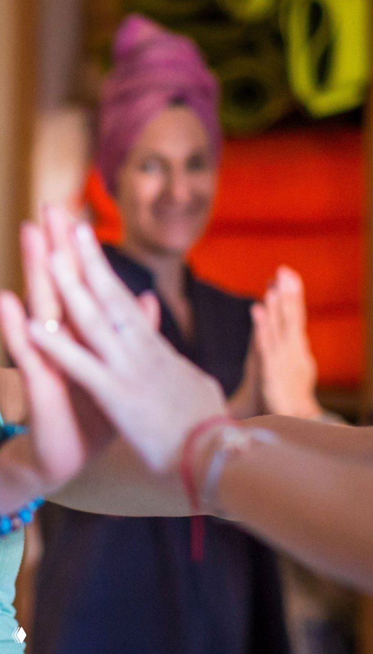 Руки и ладони участниц соединяются в жесте поддержки на занятии по движению; на заднем плане женщина-инструктор наблюдает и улыбается.