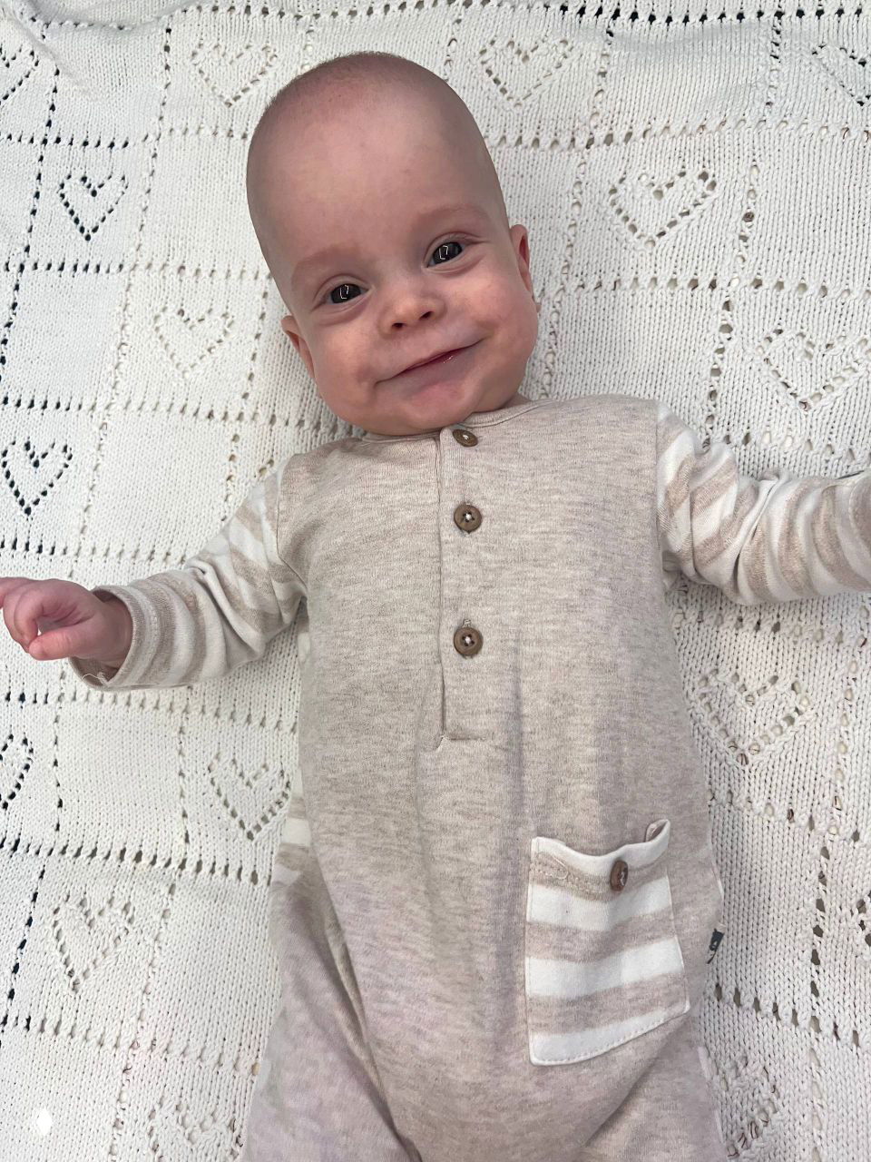 Улыбающийся Ваня лежит на вязаном пледе в светлом комбинезоне; эмоциональный портрет малыша спустя месяцы после преждевременных родов, полон радости и силы.