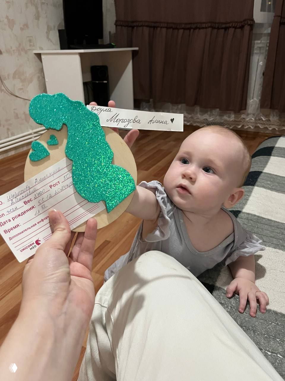 Малыш тянется к бумажной медали с надписью и подписью «Доула Морозова Алина» — символ первого прикосновения и взаимодействия с подопечной семьёй.