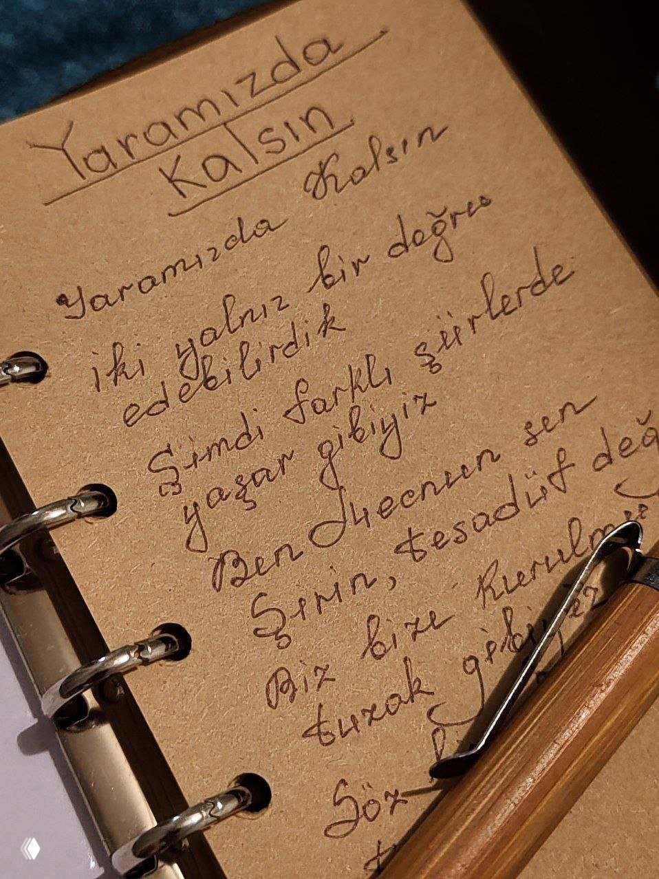 Фото разворота блокнота с аккуратно написанным текстом на турецком ("Yaramızda kalsın"), рядом видна ручка и металлические кольца блокнота, тёплый свет.