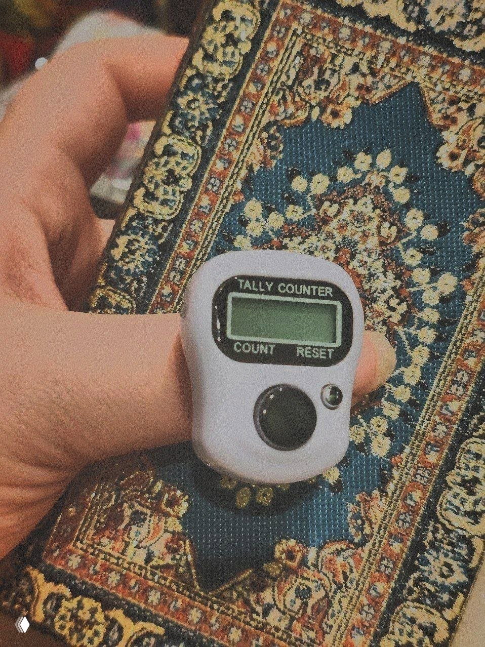 Крупный план: пальцы держат электронный счётчик (tally counter) поверх узорчатой обложки книги/блокнота — видно текстуру и прибор в кадре.
