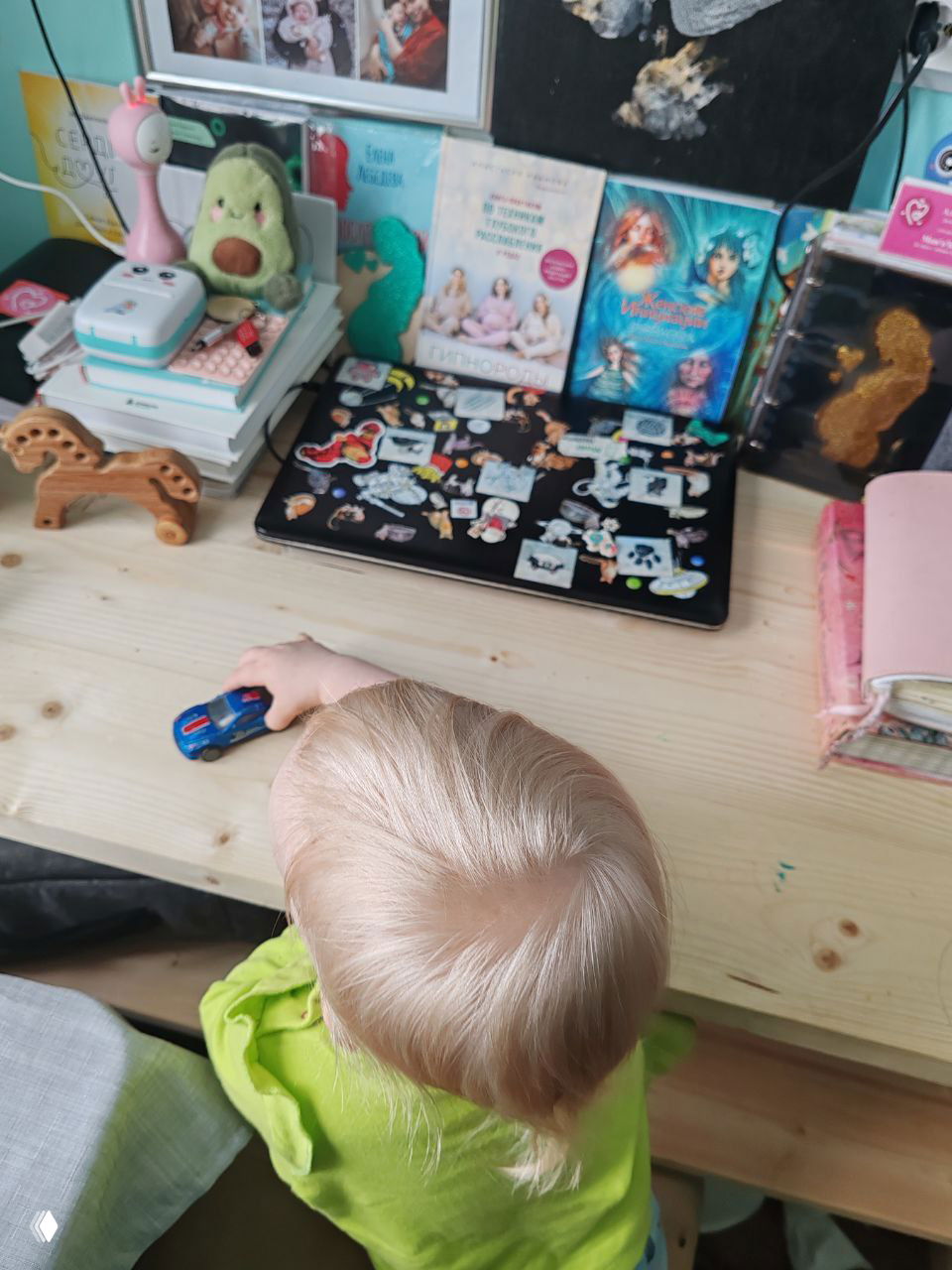 Сзади-вид ребёнка за столом в салатовой кофте, держит игрушечную машинку; на столе ноутбук с наклейками, книги и детские вещи.