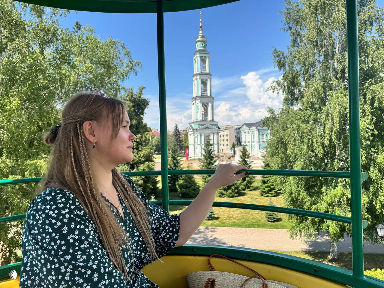 Женщина в беседке смотрит на высокий колокольный звон и купола вдалеке, зелёный парк вокруг и ясное синее небо — видовая фотография из прогулки.