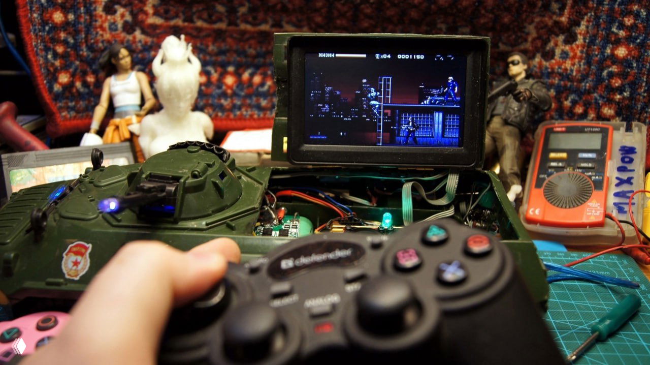 Ретро-игровая сборка в корпусе танка: прицел на верхней палубе, в переднем плане держат геймпад, на экране запущена классическая платформер-игра, фон мастерской.