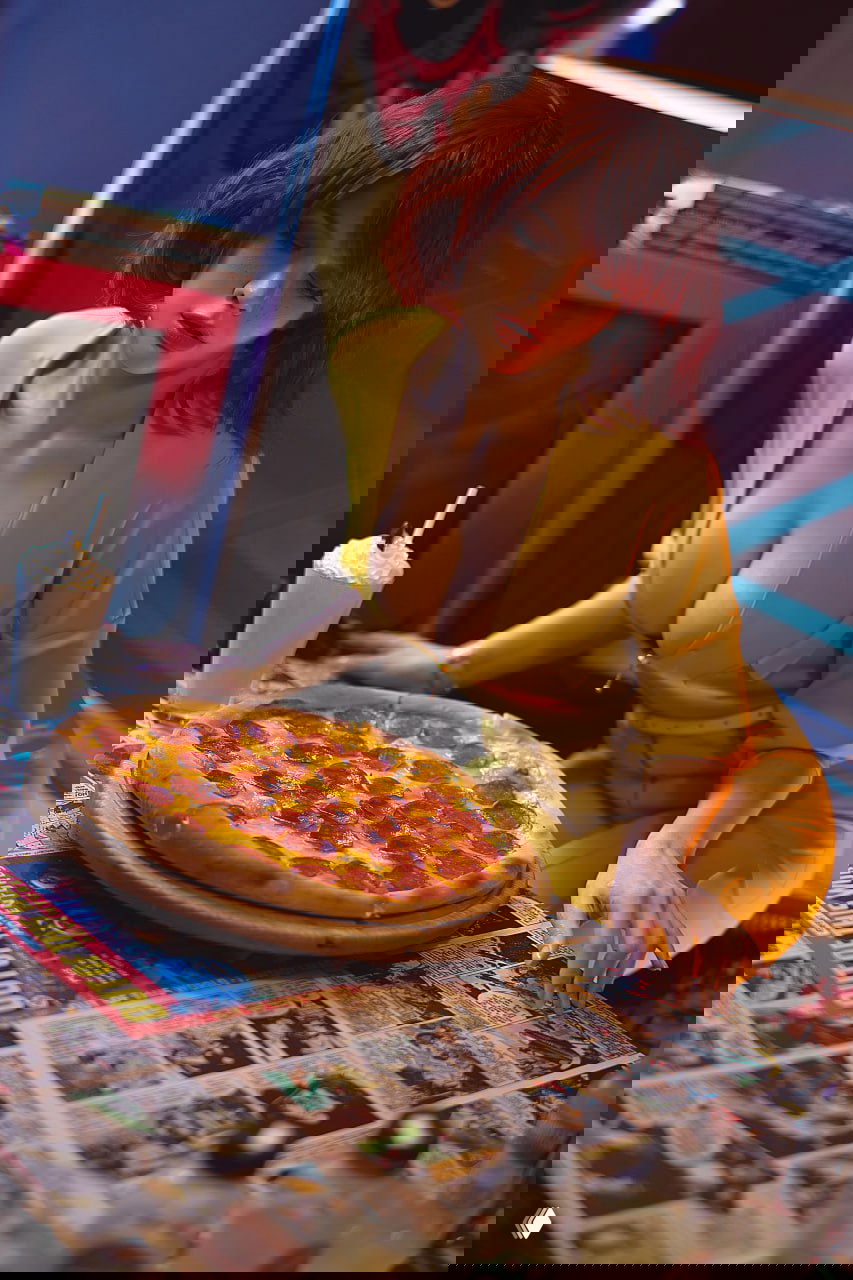 Косплей Эйприл О'Нил за столом с пиццей и милкшейком: жёлтый комбинезон, декор с комиксами и ретро‑атмосфера, художественное освещение подчёркивает образ.