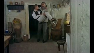 Кадр из фильма «Знахарь» (1981): мужчина несёт другого в деревенской избе, видна простая мебель и утварь.