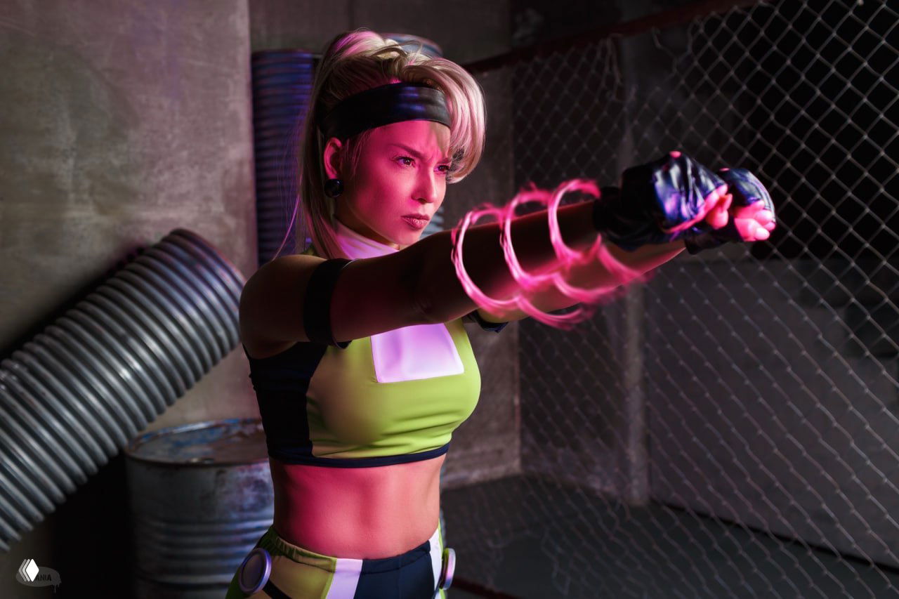 Косплеер в зеленом костюме Сони Блейд делает боевой выпад с эффектами света, сцена в сетчатой промышленной декорации.