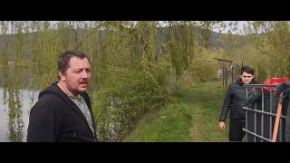 Кадр с места зарыбления: мужчина у берега, на заднем плане контейнеры и прибрежная растительность, общий план процесса зарыбления.