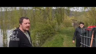 Двое мужчин у прибрежной дорожки пруда, один смотрит в камеру; видны прибрежная трава, деревья и оборудование для зарыбления рыбы в баке.