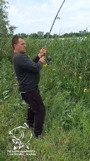Мужчина в тёмной толстовке на берегу озера держит удочку в момент подсечки рыбы, видны камыши и вода на заднем плане.