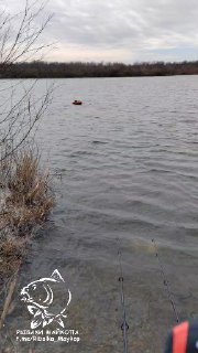 Фото Морозовских прудов в пасмурную погоду: берег с низкой растительностью, волны, оранжевый буй на воде и видимые удочки у берега.