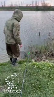 Рыбак в тёплой одежде стоит на берегу водоёма рядом с удочкой; видна прибрежная растительность и столбики на фоне воды.