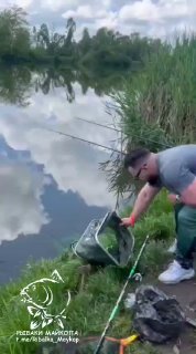 Двое рыбаков на берегу вытаскивают подсак и укладывают улов на мат; на воде отражается небо, видны удилища и снаряжение.