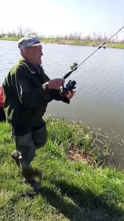 Мужчина в камуфляже стоит у кромки воды и держит катушку с удилищем во время поклёвки на берегу Утятника.
