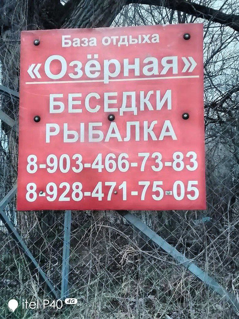 Красная табличка базы отдыха «Озёрная» с надписью «БЕСЕДКИ РЫБАЛКА» и двумя номерами телефонов на фоне кустов и деревьев