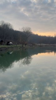 Пруд на закате с зеркальной водой, отражением неба и деревьев на берегу, вид на домики и прибрежную растительность — ранняя весна.