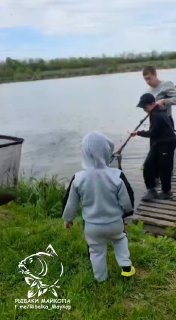 Группа рыбаков у берега платника: мужчина с подсаком достаёт улов, рядом ребёнок в светлой толстовке, видна трава и водная гладь.
