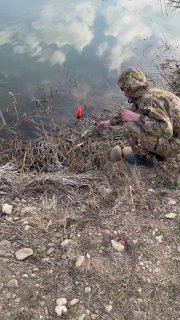 Рыбак в маскировочной одежде у воды устраивает прикормку или освобождает крючок, рядом яркий поплавок и каменистый берег.