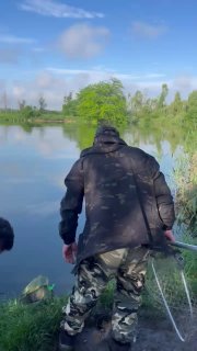 Рыбак в тёмной куртке стоит лицом к водоёму Утятник на зелёном берегу, вид со спины, спокойная водная гладь на фоне.