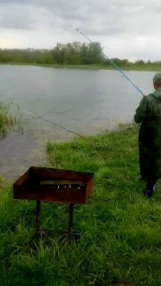 Два рыбака на фоне широкого зеркала водоёма; один в плаще держит удочку, виден мангал на переднем плане.