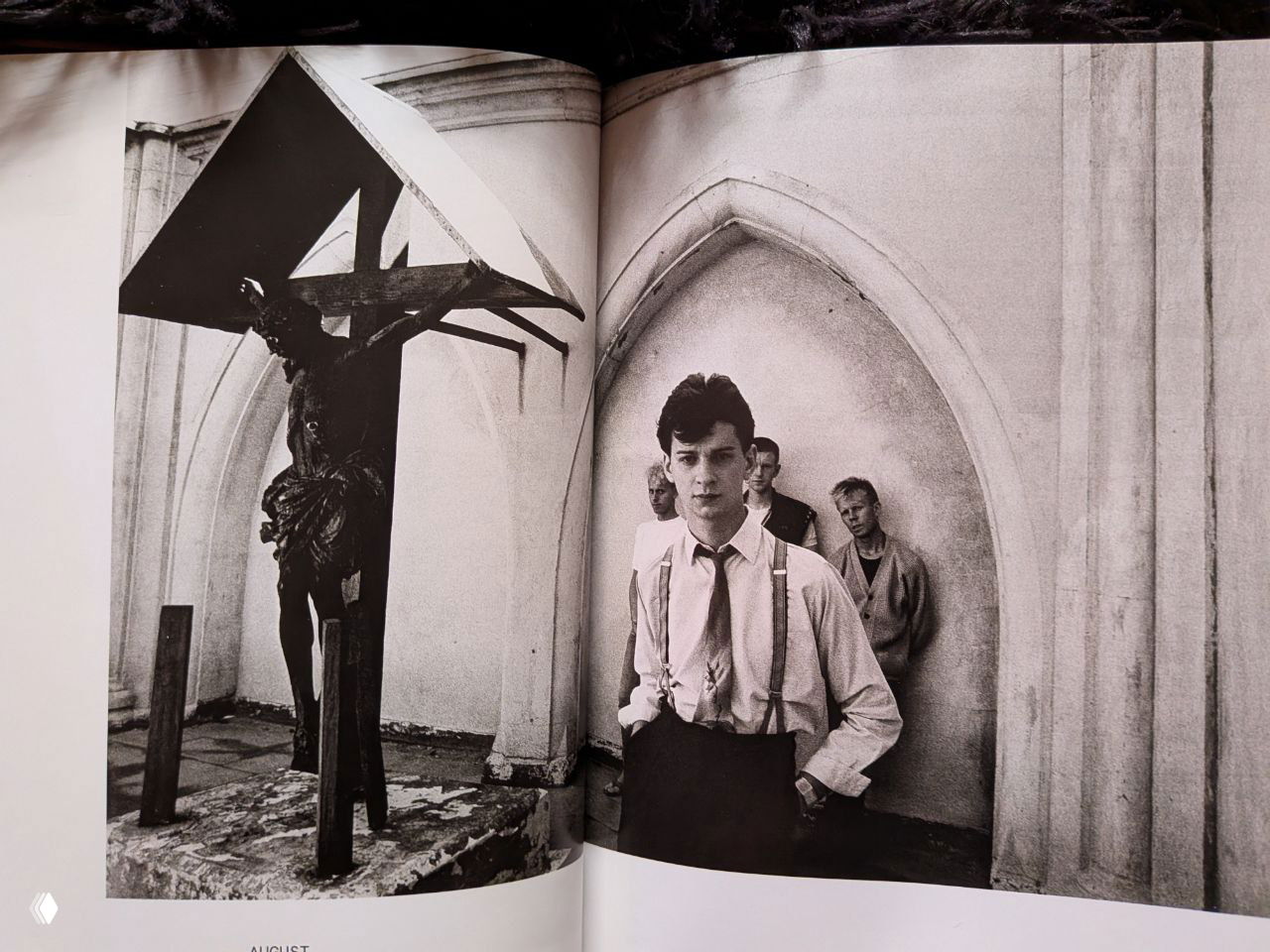 Чёрно-белая фотография из книги: молодой участник группы на переднем плане на фоне архитектурной ниши и фигуры, напоминающей распятие, мрачная готическая атмосфера.