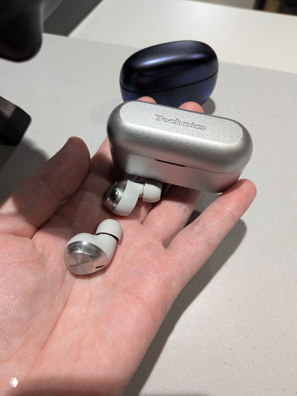 Ещё один крупный план серебристого футляра и пары наушников на ладони — подчёркнуты текстура корпуса и габариты наушников.