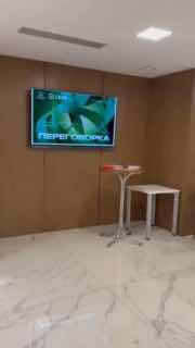 Пустой угол конференц-зала с экраном, демонстрирующим баннер «ПЕРЕГОВОРКА», столик для нетворкинга и белый мраморный пол в зале.