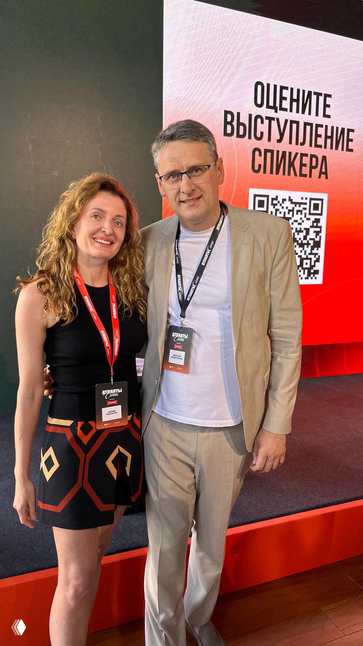Юлия Макаева фотографируется с другим спикером на фоне сцены с призывом оценить выступление спикера и QR-кодом, деловой формат события.