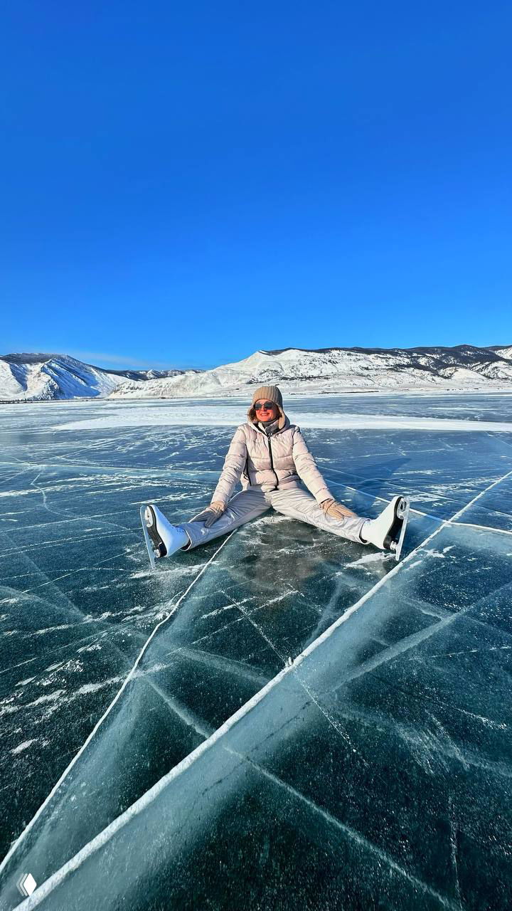 Автор в светлой куртке сидит на прозрачном льду Байкала в коньках: видна сеть трещин и глубокий синий горизонт, ощущение пространства.