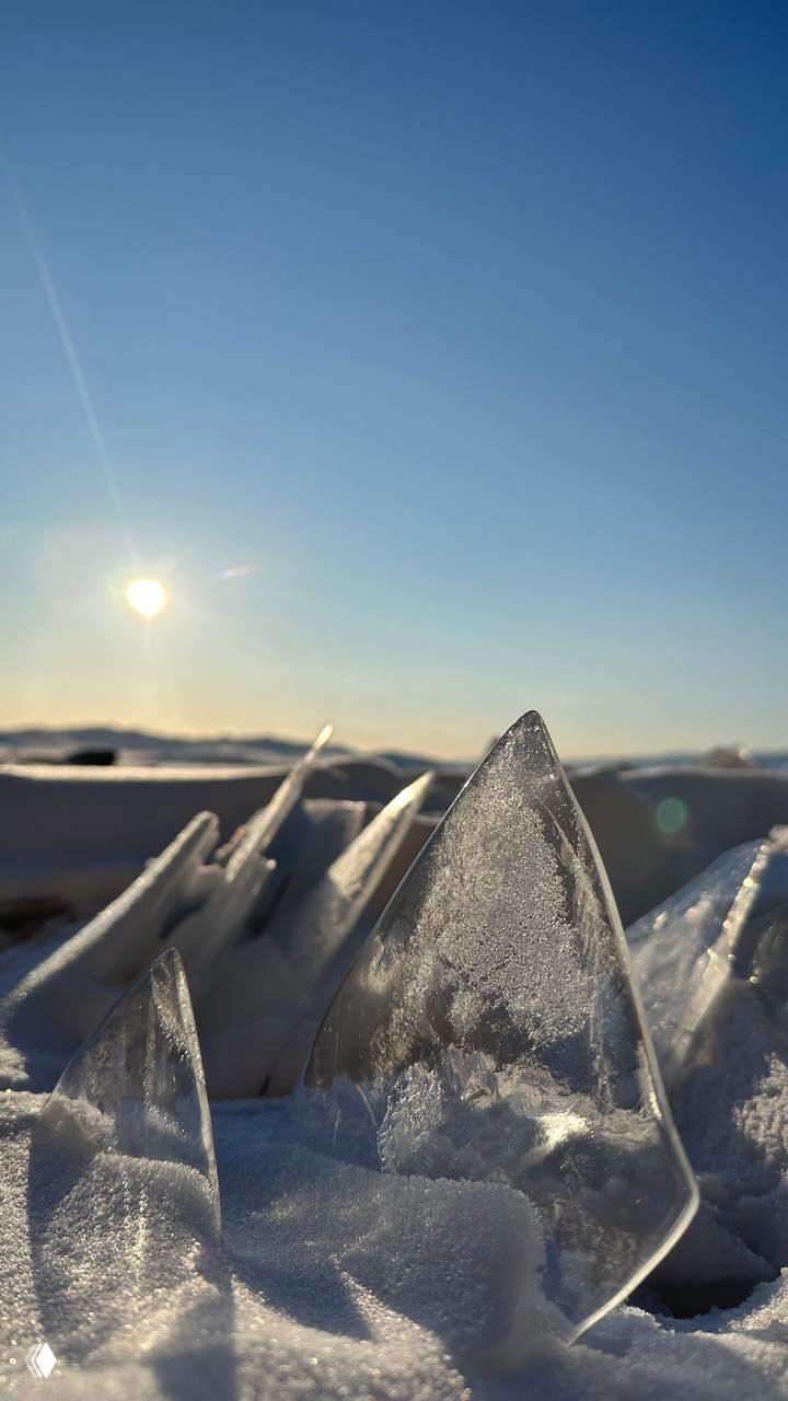Макросъёмка острых кусков прозрачного льда, торчащих из снега, на фоне солнца и горизонта — текстура и свет в деталях льда.