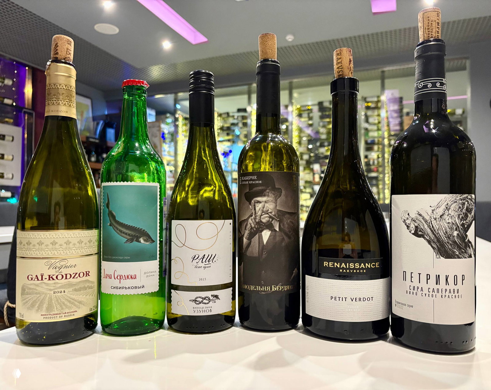 Несколько бутылок российских вин в ряд: Гай‑Кодзор, Дача Сердюка, Узунов, Раевское, Петрикор; витрина магазина видна на заднем плане.