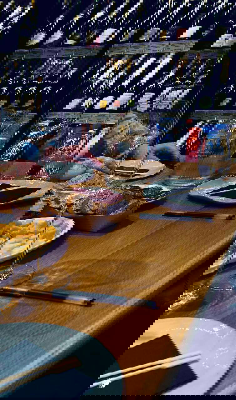 Стол дегустации с бокалами, тарелками, закусками и блокнотами для записей; на заднем плане полки с витриной винных бутылок.